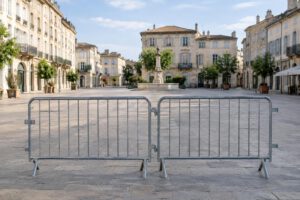 Symbolische Illustration einer menschenleeren französischen Stadtstraße mit hellen Steinfassaden: Im Vordergrund stehen mobile Absperrgitter, dahinter öffnet sich ein weiter Platz. Keine Schrift, Logos oder erkennbaren Personen; die Darstellung ist a