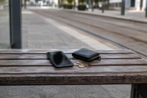 Auf einer schlichten Haltestellenbank liegen ein Smartphone mit dunklem Display und ein kleines Portemonnaie neben wenigen Münzen. Im unscharfen Hintergrund verlaufen Straßenbahnschienen; die symbolische Aufnahme zeigt keine Personen, Schrift oder Ma