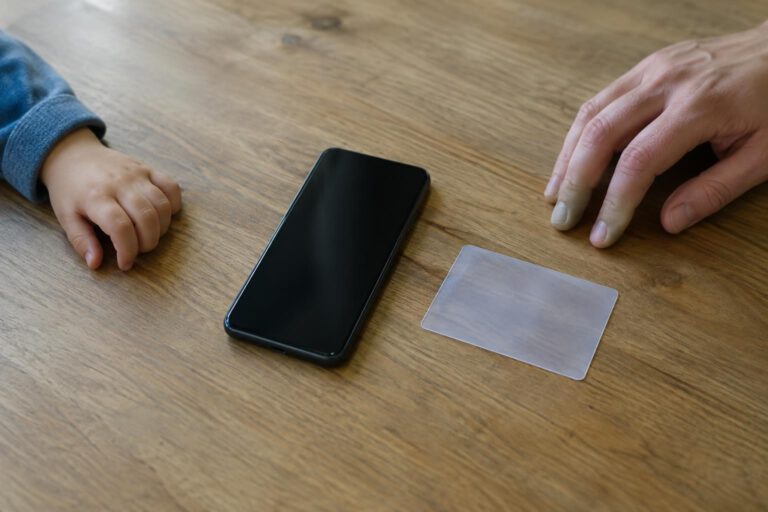 Ein Smartphone mit dunklem Display liegt neben einer durchscheinenden, völlig unbeschrifteten Karte auf einem Küchentisch. Eine kleine und eine große Hand ruhen mit Abstand daneben; die reduzierte, illustrative Inszenierung zeigt weder Gesichter noch